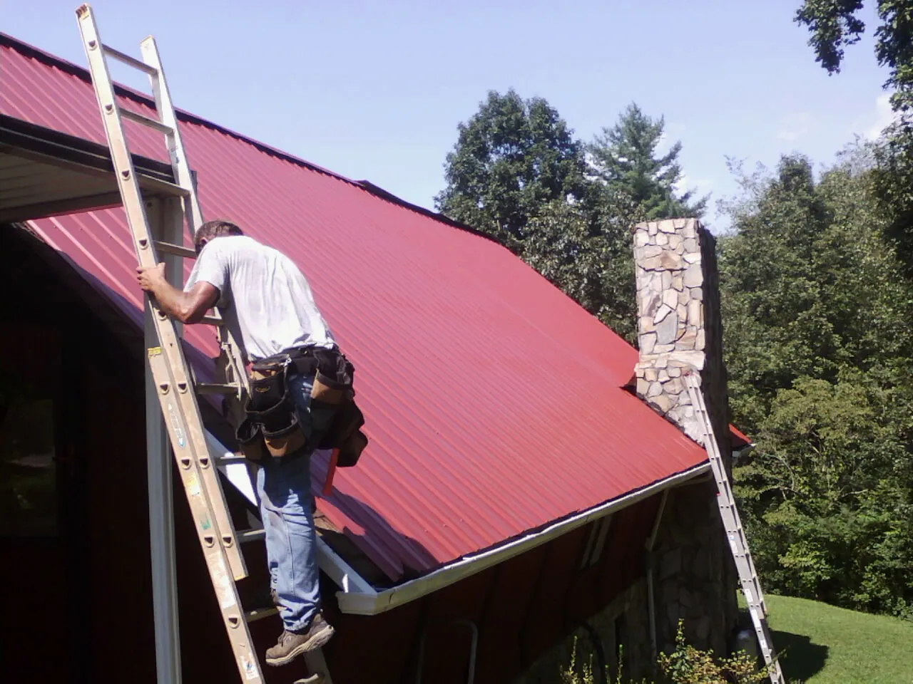 Roofer on a ladder beside a red metal roof and stone chimney — Soffit & Fascia Repair by Roofing Brothers in Blount